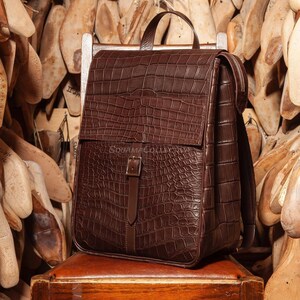 May include: A brown leather backpack with a crocodile skin effect. The backpack features a top handle, adjustable shoulder straps, and a front flap with a buckle fastening. The bag is on a wooden surface.