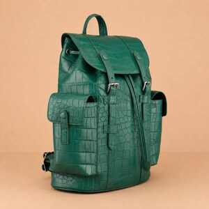 May include: A deep emerald green backpack featuring a textured crocodile-embossed pattern. The bag includes a top handle, a fold-over flap with two vertical buckled straps, a drawstring closure, and two side pockets with snap-button closures.