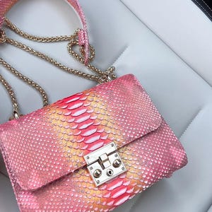 Może przedstawiać: Różowa i żółta torebka z wzorem skóry węża ze złotym łańcuszkiem. Prostokątna torba ma srebrne zapięcie. Kolor torby przechodzi od różowego do żółtego i czerwonego, tworząc efekt gradientu.