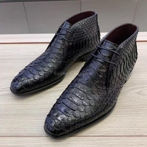 May include: A pair of black leather ankle boots with a textured, scale-like pattern. The boots have laces and a low heel. The interior is a dark burgundy color. The boots are displayed on a light wood surface.