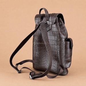 Pode incluir: Uma mochila castanho-escura com um padrão texturizado de pele de crocodilo. A mala inclui uma pega superior, alças ajustáveis, uma aba com fecho seguro e um bolso lateral. O design apresenta uma silhueta estruturada.