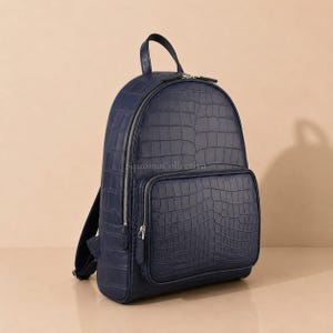 Pode incluir: Uma mochila azul-marinho com um padrão de textura de pele de crocodilo. A bolsa inclui um compartimento principal grande com fecho de correr prateado, um bolso frontal menor com fecho a condizer, uma alça superior e alças ajustáveis.