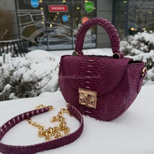 May include: A deep purple handbag with a textured snakeskin pattern, a top handle, and a gold-colored clasp. The bag includes a detachable strap with a gold chain. The bag is sitting on a snowy surface.