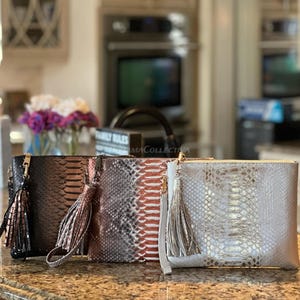 以下が含まれることがあります： ヘビ革模様の長方形クラッチバッグ4つのコレクション。バッグは黒、ブロンズ、赤のアクセントが入ったグレー、シルバーです。各バッグにはお揃いのタッセルとゴールドカラーのジッパーが付いています。