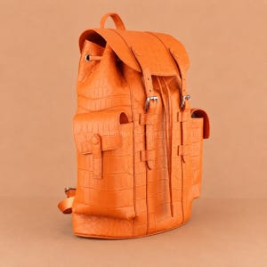 Op de afbeelding: Een feloranje rugzak met een gestructureerd krokodillenleerpatroon. De tas heeft een bovenflap met twee verticale riemen, zilverkleurige metalen gespen, een trekkoordsluiting en twee zijzakken met drukknopen aan de voorzijde.