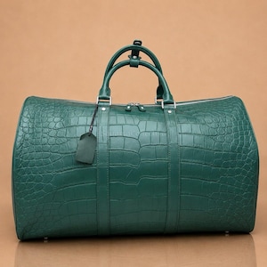 Peut inclure: Un sac de voyage vert foncé avec un motif texturé en peau de crocodile. Le sac comprend deux poignées supérieures, une fermeture éclair argentée et une étiquette de bagage en cuir vert assortie attachée par un cordon noir fin.