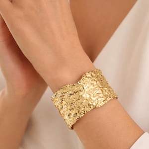 May include: A wide, textured gold cuff bracelet. The bracelet has an organic, rippled surface. The jewelry is worn on a wrist, with a white shirt visible in the background. The cuff is a statement piece.
