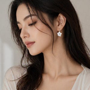 Puede incluir: Primer plano de una persona con pendientes de flores plateadas. Los pendientes tienen un pequeño diseño de flor blanca. La persona tiene el pelo oscuro y lleva una blusa de color claro. Los pendientes son una joya delicada.
