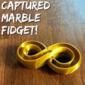 May include: A gold-coloured infinity-shaped fidget toy with a small white marble captured inside the track. The text "CAPTURED MARBLE FIDGET!" is printed in white letters above the object. The toy features a continuous loop design.