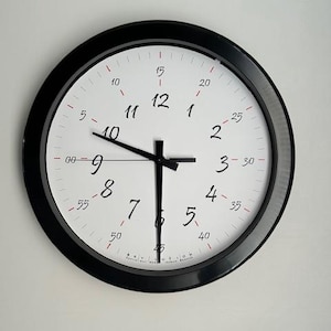 May include: A round wall clock with a black frame and a white face. The clock features black numbers and hands, with red minute markers. The time is approximately 9:30. The clock is mounted on a white wall.