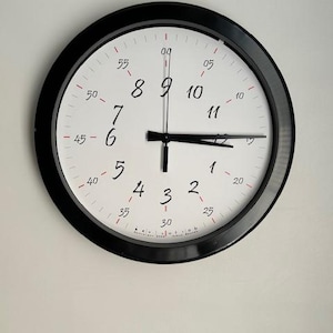 May include: Round black wall clock with a white face. The clock features black hands and black numbers. Red minute markers are around the edge of the clock face. The time is approximately 3:15.