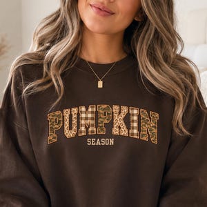 May include: A dark brown crewneck sweatshirt featuring the text PUMPKIN SEASON in a collegiate font. The letters of the word PUMPKIN are filled with various patterns including leopard print, camouflage, and plaid, while the word SEASON is plain.