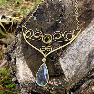 Peut inclure: Un collier doré avec un pendentif en pierre précieuse bleue irisée en forme de goutte d'eau. Le collier présente un motif décoratif en spirale et est présenté sur une surface en bois vieilli, avec un arrière-plan extérieur naturel.