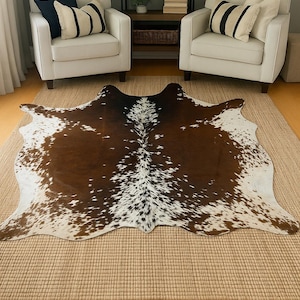 May include: A brown and white cowhide rug with a unique spotted pattern. The rug is placed on a woven jute rug, with two white armchairs and a bookshelf in the background.