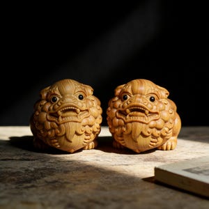 Può includere: Due statue di leoni guardiani in legno intagliato in modo intricato. Le statue sono marrone chiaro con dettagli, tra cui occhi espressivi e un'espressione feroce. I leoni sono seduti e hanno una criniera e un corpo strutturati.