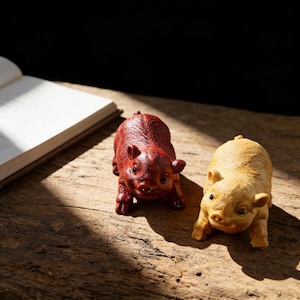 Pode incluir: Duas pequenas figuras de porco. Uma é de cor vermelho escuro e a outra é bege claro. Ambos os porcos estão virados para a frente com as pernas abertas. As figuras estão sobre uma superfície de madeira, com um livro ao fundo.