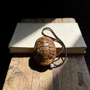 Puede incluir: Un colgante de tortuga de madera detallado con un cordón marrón y una pequeña cuenta blanca. El colgante de tortuga está tallado con un patrón de caparazón texturizado. El colgante descansa sobre una superficie de madera junto a un libro.