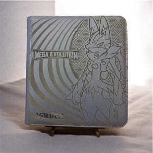 May include: A silver-toned trading card binder featuring a line-art illustration of the character Lucario. The cover displays the text MEGA EVOLUTION and the brand name vaultx in the lower left corner, with a geometric pattern background.