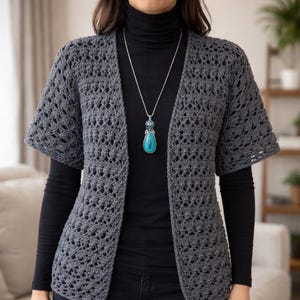 Puede incluir: Un cárdigan de crochet gris con frente abierto y mangas cortas. El cárdigan tiene un diseño de tejido abierto y texturizado. Un collar de plata con un colgante turquesa se usa sobre un cuello alto negro.