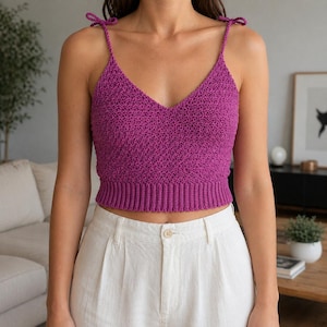 May include: A magenta crochet top with a V-neck and tie straps. The top has a ribbed bottom band and a textured stitch pattern. The model is wearing the top with white pants.