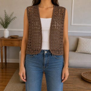 Può includere: Un gilet senza maniche all'uncinetto marrone con bordo smerlato. Il gilet aperto è indossato sopra una canotta bianca e jeans blu. Il design a trama aperta del gilet è visibile. Lo stile generale è casual e rilassato.
