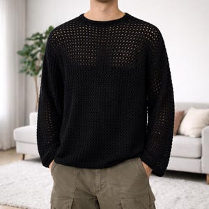 Peut inclure: Un pull noir à mailles ajourées, avec un col rond et des manches longues. Le pull présente un motif texturé en forme de grille. La personne porte un short cargo vert olive. L'arrière-plan comprend un canapé blanc, des coussins et une plante.