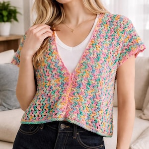 Könnte beinhalten: Ein farbenfroher, kurzärmeliger Häkelcardigan in Pastelltönen von Rosa, Blau, Gelb und Grün. Der Cardigan mit offenem Front hat einen V-Ausschnitt und wird über einem weißen Oberteil getragen. Der Cardigan wird mit dunkelblauen Jeans kombiniert.
