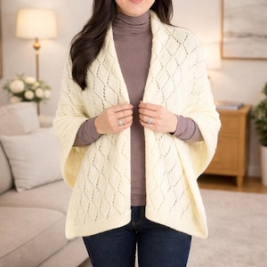 May include: Cream-coloured knitted shawl with a diamond pattern. The shawl is draped over the shoulders. The person is wearing a brown turtleneck and blue jeans. The shawl is a cosy and stylish accessory.