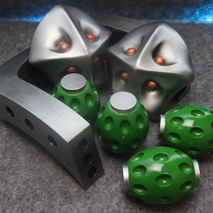 Può includere: Una collezione di oggetti di scena futuristici, tra cui due copricapi geometrici argentati con dettagli arancioni, diversi oggetti sferici verdi e un pezzo curvo grigio metallizzato con ritagli circolari.