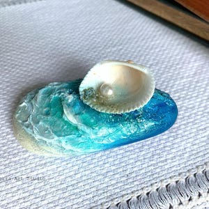 May include: A handcrafted, ocean-themed decorative piece. It features a blue and turquoise painted stone with a seashell and a pearl accent. The seashell is white and beige, and the stone has a textured, wave-like appearance. The piece is set on a white textured surface.