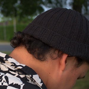 May include: A black knit beanie with a ribbed texture and a folded cuff, worn on the head. The beanie features a consistent vertical knit pattern throughout the crown and the brim, providing a snug fit around the upper portion of the head.