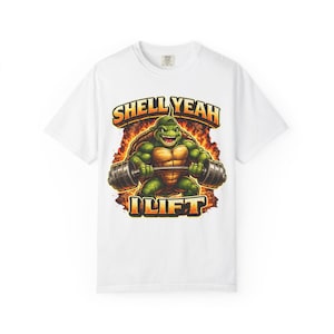 Op de afbeelding: Wit t-shirt met een cartoon schildpad die gewichten heft. De schildpad is groen en bruin, met de woorden "SHELL YEAH I LIFT" in oranje en geel. Het ontwerp heeft een vurige achtergrond.