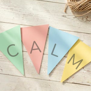 May include: A banner with the word "CALM" spelled out across four triangular flags. The flags are pastel colors: green, pink, blue, and yellow. The letters are gray. The banner is on a light wood surface with a ball of twine in the background.