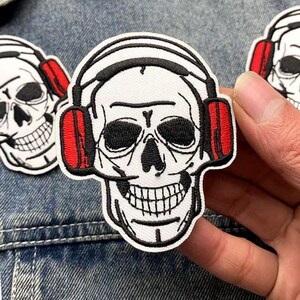 Puede incluir: Parche bordado con una calavera blanca que lleva auriculares negros con orejeras rojas. La calavera tiene ojos negros y una amplia sonrisa. El parche está sobre un fondo de mezclilla.