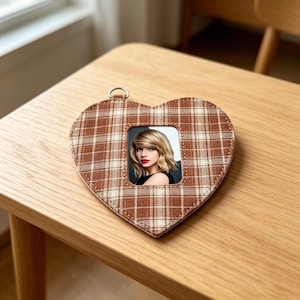 May include: Heart-shaped photo frame with a brown and cream plaid pattern. The frame has a small metal loop at the top and holds a colour photograph. The frame is sitting on a wooden surface.