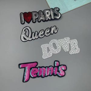 Peut inclure: Plusieurs écussons à paillettes avec des motifs de texte. Les écussons incluent "I ❤️ PARIS" avec un cœur rouge, "Queen", "LOVE" et "Tennis". Chaque écusson a une bordure noire et est posé sur un fond gris.