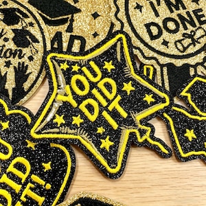 Op de afbeelding: Een verzameling zwarte en gouden glitterpatches. De centrale patch is stervormig met de gele tekst "YOU DID IT". Andere patches bevatten afstudeerthema's en de tekst "I'M DONE".
