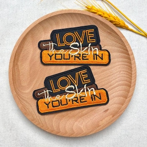 May include: Two embroidered patches with the text "LOVE the Skin YOU'RE IN" in white and gold lettering on a black and orange background. The patches are displayed on a wooden plate.