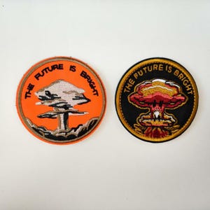 Puede incluir: Dos parches bordados circulares. Uno es naranja con un diseño estilizado de nube de hongo y el texto "THE FUTURE IS BRIGHT". El otro es negro con un diseño de nube de hongo rojo y amarillo y el mismo texto.