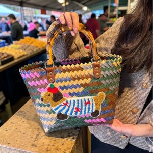 Peut inclure: Un sac fourre-tout tissé avec une poignée en bambou et des détails en cuir. Le sac présente un motif rayé coloré et une application de teckel portant un pull rayé bleu et blanc avec des nœuds rouges. Le sac est tenu par une personne.