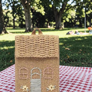 Könnte beinhalten: Hausförmige Handtasche aus geflochtenem Stroh mit Henkel. Die Tasche hat gestickte Details wie Fenster, eine Tür und Blumendesigns. Sie liegt auf einer rot-weiß karierten Picknickdecke, mit einer Parkszene im Hintergrund.