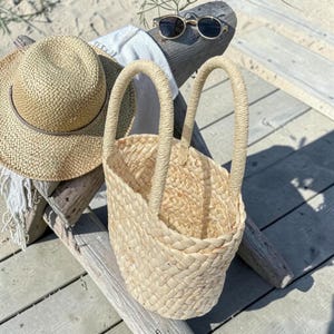 Op de afbeelding: Een geweven strooien draagtas met twee handvatten, een strohoed en een zonnebril rusten op een houten oppervlak. De tas is lichtbeige en heeft een getextureerde uitstraling. De hoed is ook strokleurig met een bruine band.