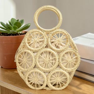 Op de afbeelding: Een beige geweven strooien handtas met een ronde handgreep. De tas heeft een uniek ontwerp met cirkelvormige geweven patronen. De binnenkant is van lichtbeige stof. De tas staat op een houten oppervlak.