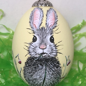 Puede incluir: Adorno de huevo de Pascua pintado a mano con la cara de un conejo gris y blanco con orejas rosas y nariz rosa. El huevo es de color amarillo pálido, con una tapa plateada y una cuerda para colgar. El conejo está rodeado de hierba verde y bayas rojas.