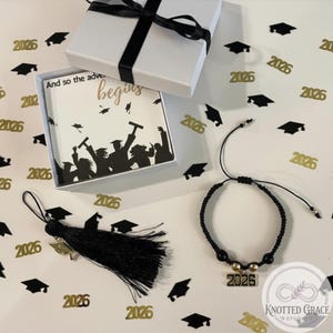 Puede incluir: Un juego de regalo con temática de graduación que presenta una caja blanca con una cinta negra, una borla negra y una pulsera negra con un dije "2026". La caja contiene una tarjeta con el texto "And so the adventure begins". Confeti dorado y negro con forma de gorro de graduación.