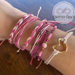 May include: A collection of pink and white braided and beaded bracelets. One bracelet features a gold heart charm. The bracelets are displayed on a wrist. The image includes the text "Knotted Grace Studio" in a circular logo.