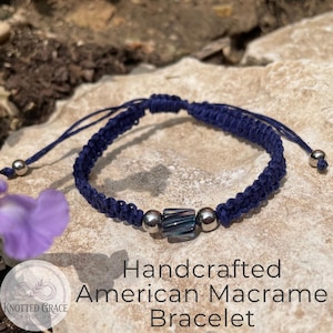 May include: A handcrafted navy blue macrame bracelet with a central rectangular bead and two silver beads. The bracelet is adjustable with sliding knots and is displayed on a natural stone surface. The text "Handcrafted American Macrame Bracelet" is visible.
