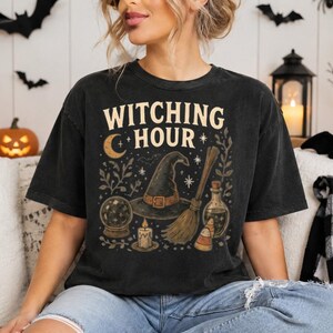 May include: A black short-sleeve t-shirt featuring a graphic print with the text "WITCHING HOUR" in cream letters. The design includes a witch hat, a broomstick, a crystal ball, a lit candle, a potion bottle, candy corn, and a crescent moon.