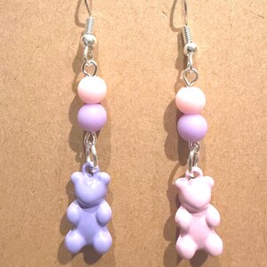 May include: A pair of dangle earrings with silver-tone hooks. Each earring features three pastel beads in pink and lavender, connected by silver-tone links. A small, lavender and pink teddy bear charm dangles at the bottom.