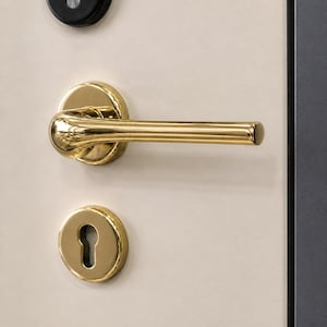 May include: Close-up of a gold-colored door handle, keyhole, and a circular plate. The handle is a lever style with a curved design. The keyhole is oval-shaped. The hardware is mounted on a cream-colored door.